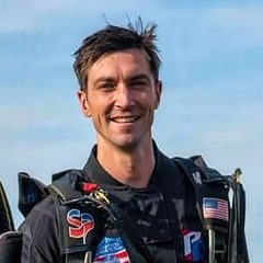 A smiling man in a black jumpsuit with various patches, including a flag, stands outside under a blue sky. He appears to be in a skydiving suit.