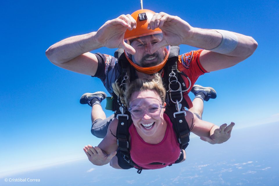 A woman is skydiving in tandem with an instructor. She is smiling widely, wearing goggles and a pink shirt, while the instructor is behind her wearing an orange helmet, making a heart shape with his hands. The sky is clear and blue.