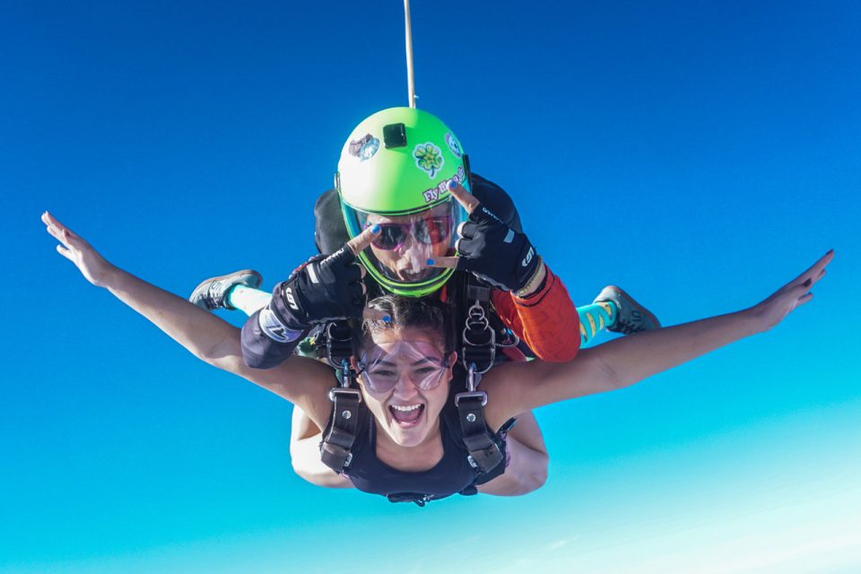 A person is tandem skydiving with an instructor. Both are wearing harnesses and goggles, smiling widely against a clear blue sky. The instructor has a green helmet, while the skydiver spreads their arms in excitement.