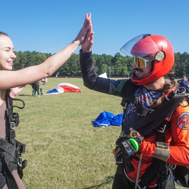 Two skydivers celebrate with a high-five after landing. The woman on the left wears a brown jumpsuit, while the person on the right wears a red jumpsuit and helmet. Green grass and parachutes are visible in the background.