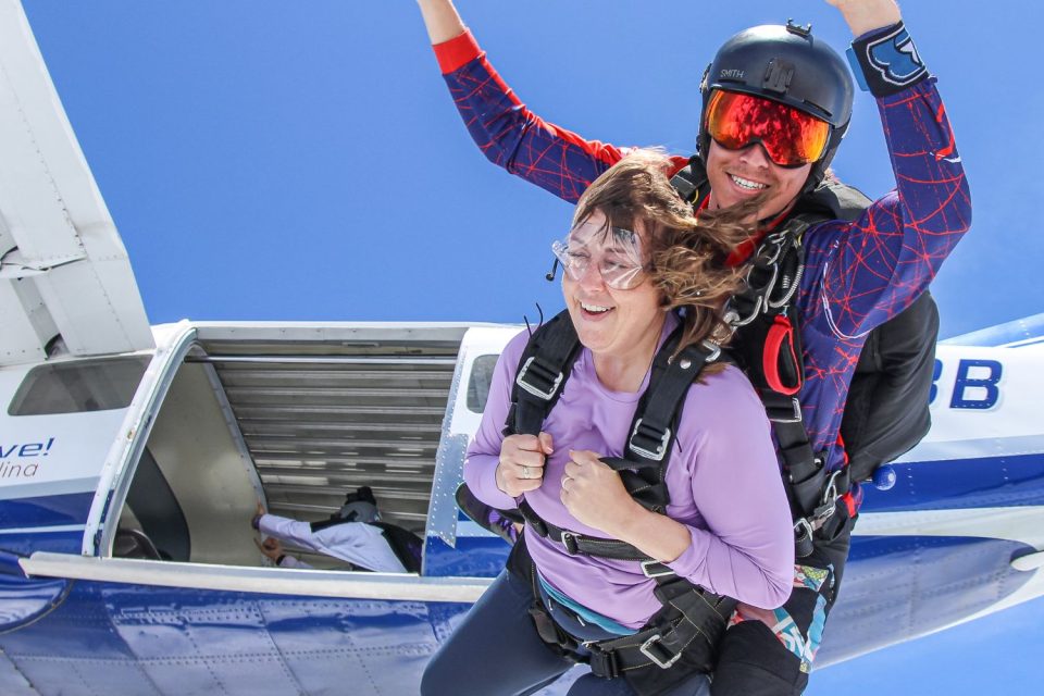 Two people tandem skydiving, exiting a plane mid-air. The instructor wears a helmet and sunglasses, guiding the other person who is smiling with arms raised. The plane is white and blue with an open side door. Clear blue sky in the background.