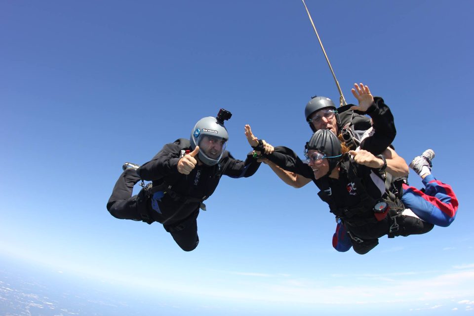 Three skydivers in freefall against a clear blue sky. One person is holding a camera, another is gesturing with a thumbs-up, and the third is smiling with arms spread wide. They are wearing helmets and gear, with a landscape visible far below.