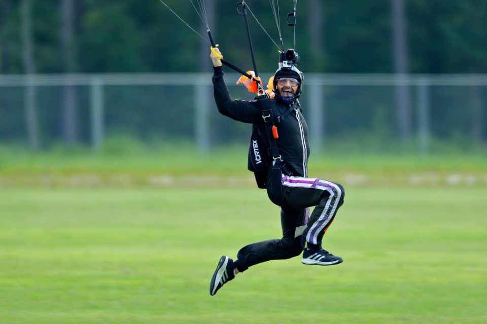 A skydiver in a black jumpsuit and helmet is landing on a grassy field with a parachute. They appear joyful, with one leg extended and arms holding the parachute strings. A blurred forest is in the background.