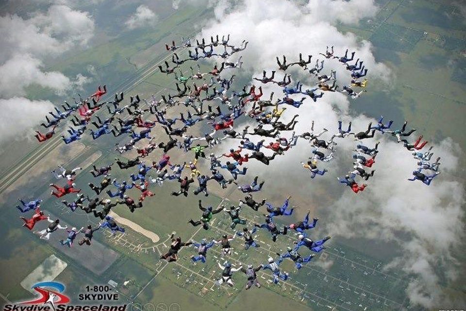 A large group of skydivers in colorful suits forms intricate patterns in the sky above a rural landscape, with clouds scattered throughout the background. The ground is visible below, showing fields and roads.