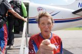 A person wearing a red and blue jumpsuit points toward the camera while standing in front of an airplane. Another person in a green and black outfit is entering the aircraft. The scene suggests skydiving activities.