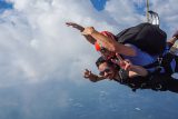 Two people tandem skydiving against a backdrop of clouds. The instructor is wearing a blue shirt and red helmet, while the other person is wearing sunglasses, making a peace sign with one hand. They appear to be enjoying the thrill of the dive.