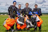 Four skydivers standing on grass, wearing black jumpsuits, holding bright orange and black parachutes. They each have helmets, two holding blue and two carrying black. The sky is partly cloudy, and they smile at the camera.