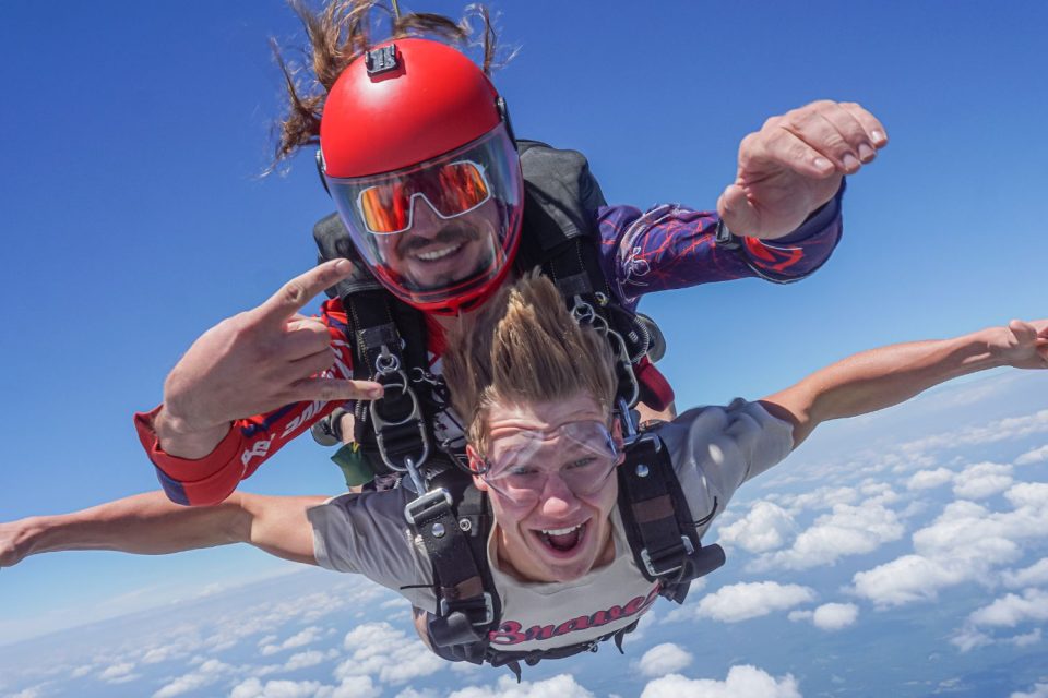 Two people skydiving in tandem with a clear blue sky and clouds below them. The front person wears goggles and a casual shirt, while the instructor behind has a red helmet and sunglasses, holding onto the other person. Both look excited.