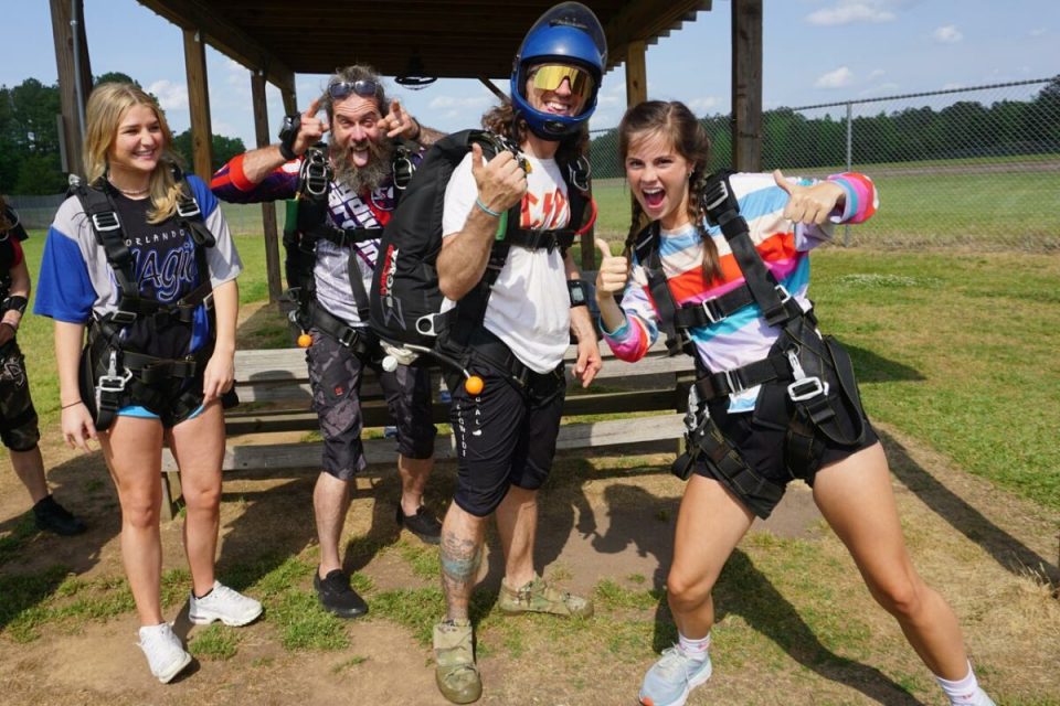 A group of four people wearing skydiving gear smile and pose enthusiastically at an outdoor skydiving center. One person is wearing a helmet and pointing thumbs up. A bench and a fence are visible in the background.