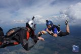 Two people in skydiving gear are free-falling through the sky, playfully passing a toy potato between them. They are surrounded by clouds and have a wide landscape below. Both are wearing helmets and jumpsuits.