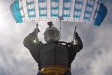 A skydiver captured mid-descent, wearing a white helmet and a dark jumpsuit, holding the straps of a blue and white parachute above. The sky is partly cloudy in the background.