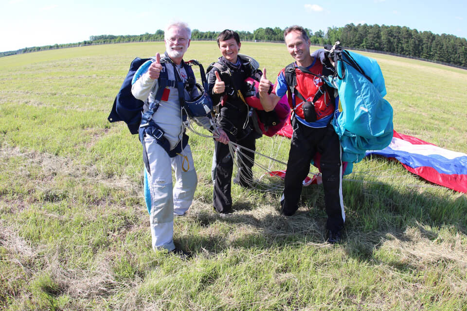 Three people in skydiving gear stand on a grassy field, smiling and giving thumbs up. Parachutes are visible on the ground behind them. Trees line the horizon under a clear blue sky.