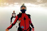 Skydivers in freefall, captured mid-air with one in the foreground wearing a red suit and helmet camera. The earth is visible upside down in the background. Other divers are seen behind, showcasing a dynamic skydive scene.