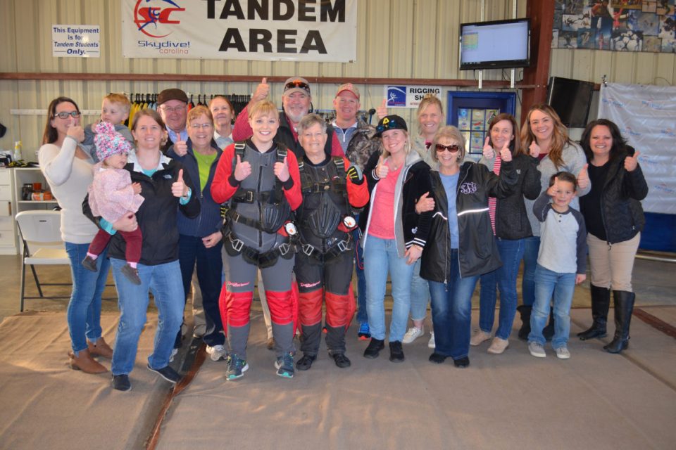 A large group of people, including men, women, and children, gather in a hangar designated for tandem skydiving. Two individuals in red jumpsuits and harnesses stand in front, while others smile and give thumbs-up gestures.