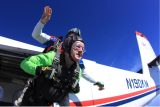 Two people are skydiving tandem from an airplane. The lead person wears a green suit and goggles, expressing excitement. The instructor behind wears a helmet and has an arm up. The plane's registration number is visible, and the sky is clear and blue.