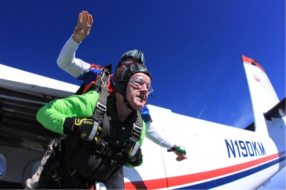 Two people are skydiving tandem from an airplane. The lead person wears a green suit and goggles, expressing excitement. The instructor behind wears a helmet and has an arm up. The plane's registration number is visible, and the sky is clear and blue.