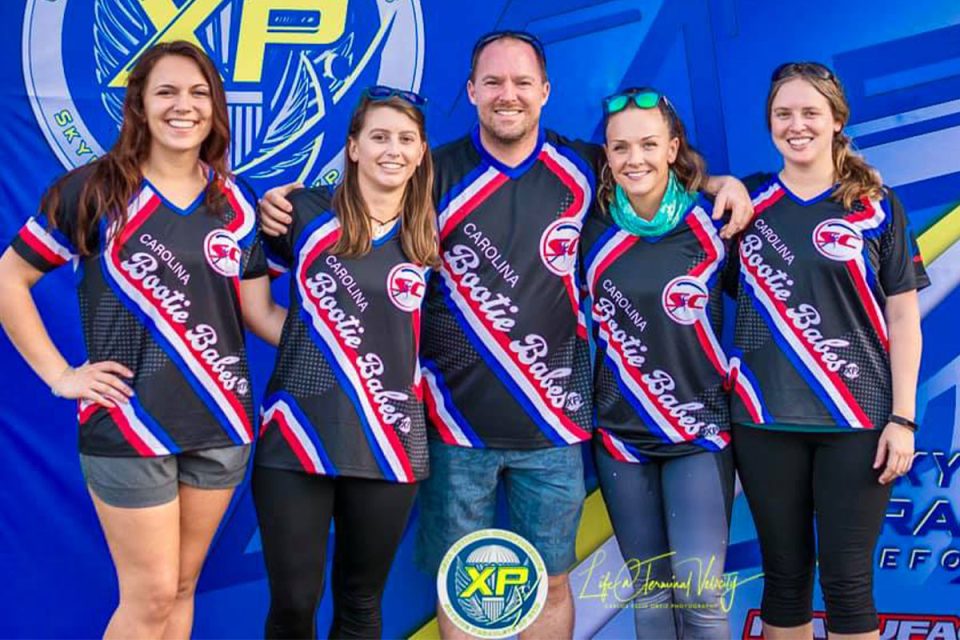 Five people are smiling and standing together in front of a blue banner with logos. They are wearing matching black, red, and blue sports jerseys with "Carolina SC Boogie Babes" written on them.