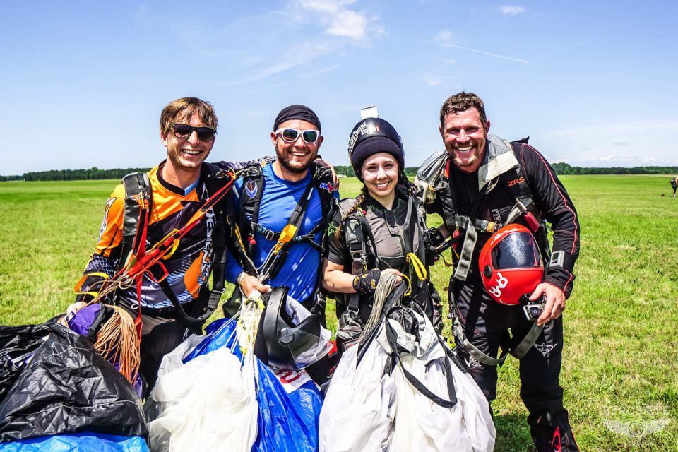 Four skydivers stand smiling on a grassy field, wearing their parachuting gear. They hold their parachutes in hand, and the sky is clear and sunny.