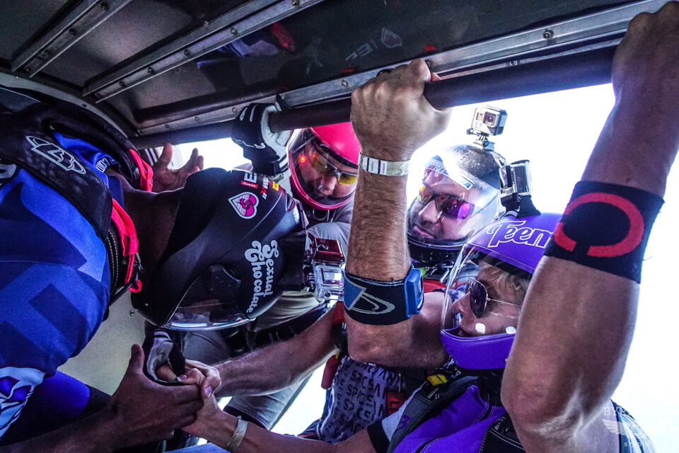 A group of four skydivers in colorful gear prepare to jump from an airplane. They are wearing helmets with visors and cameras, grasping each other's hands for support and coordination. The sky is visible through the open doorway.