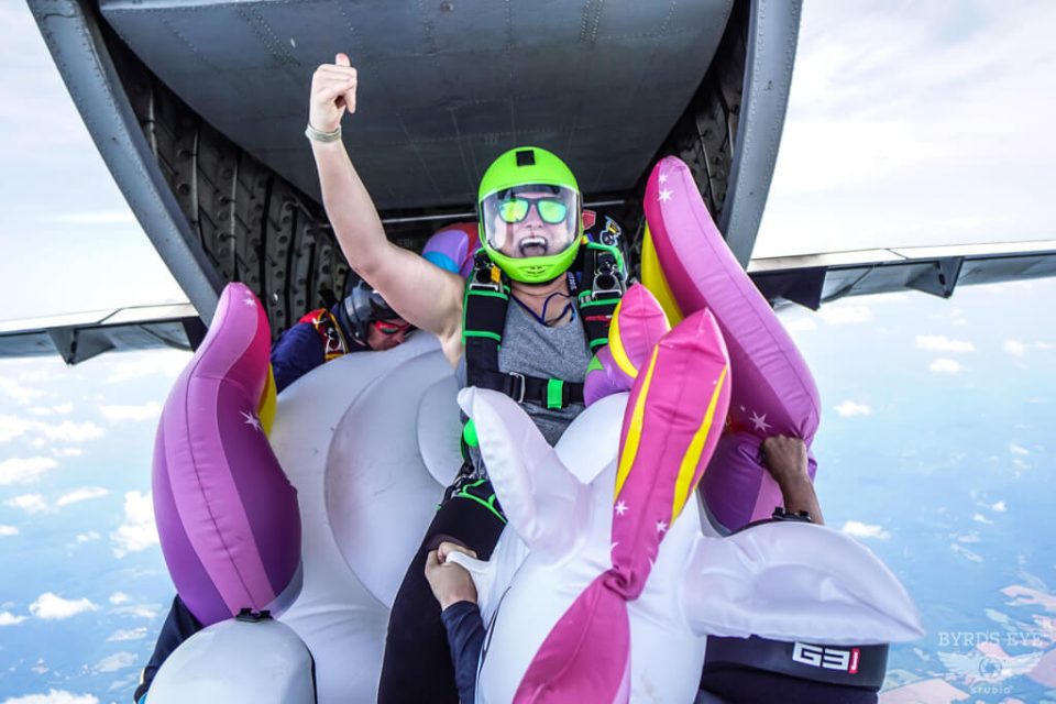 A person wearing a green helmet and sunglasses is skydiving from an aircraft, seated on an inflatable unicorn float. The sky and clouds are visible in the background. The person appears to be excited, with one arm raised.