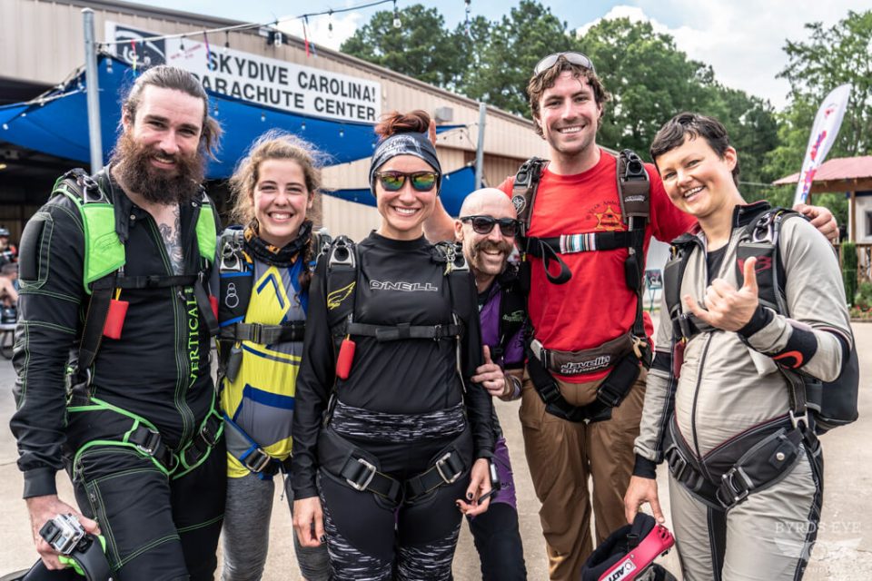 A group of six skydivers in jumpsuits and harnesses stand smiling in front of the Skydive Carolina Parachute Center. They appear happy and excited, ready for a jump. Trees and a building sign are visible in the background.