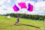 A skydiver wearing a purple jumpsuit and helmet lands on a grassy field with a pink and purple parachute. Trees and a cloudy sky are in the background.