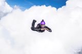 A person in a black jumpsuit and purple helmet is skydiving through a bright, cloud-filled sky. They are smiling with arms bent and hands together, surrounded by fluffy white clouds and blue sky.