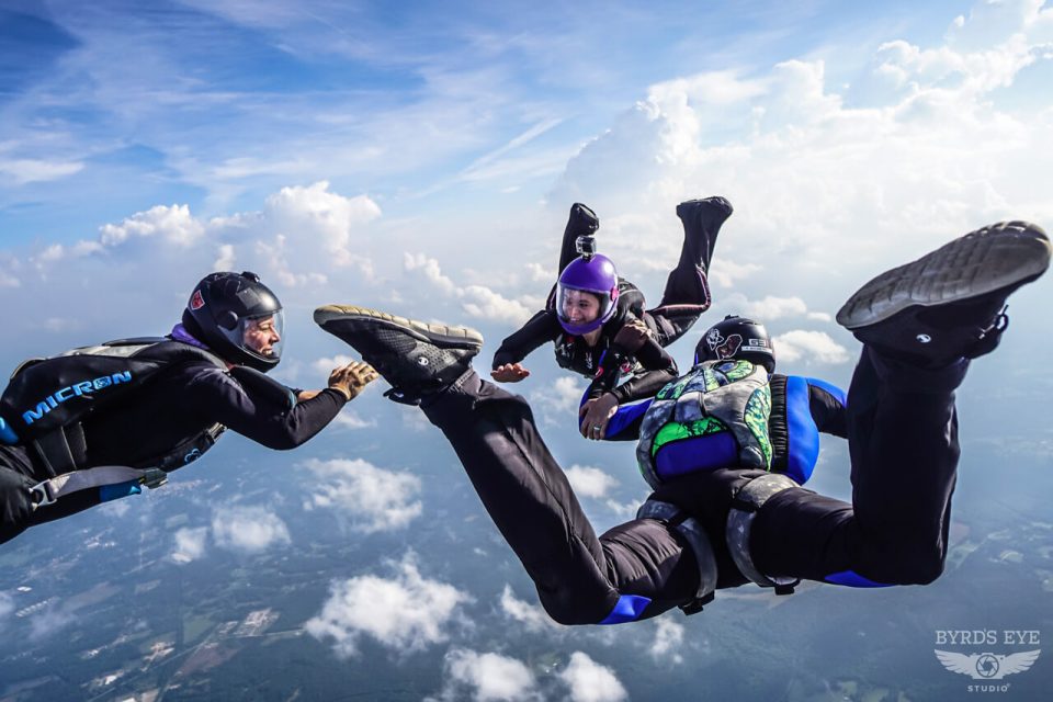 Three skydivers in mid-air, wearing black and blue jumpsuits with helmets, engage in a formation against a backdrop of a bright blue sky with scattered clouds. One diver wears a purple helmet, adding contrast to the scene.