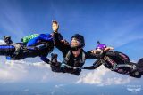 Three skydivers in mid-air formation, wearing helmets and jumpsuits, are linked by their arms against a clear blue sky with clouds. Their expressions convey focus and excitement during the freefall.