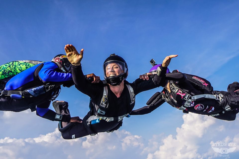 Three skydivers in mid-air, smiling and holding hands against a backdrop of blue sky and clouds. They wear helmets and gear, creating a sense of motion and camaraderie as they freefall together.