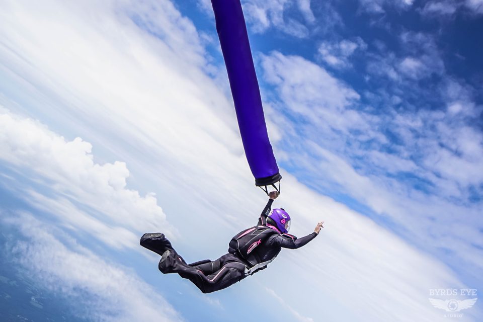 A skydiver in a black jumpsuit and purple helmet is gripping a long purple fabric in mid-air against a backdrop of blue sky and clouds. The skydiver appears to be performing an acrobatic maneuver.