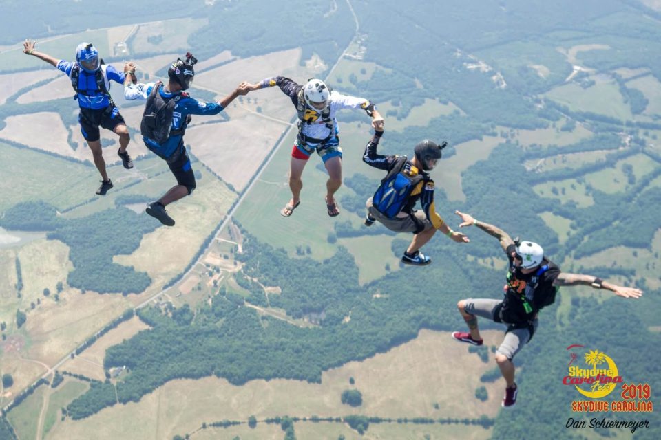 A group of five skydivers in mid-air, holding hands in a formation over a landscape of fields and trees. They are wearing helmets and parachute gear, with clear skies and distant hills in the background.