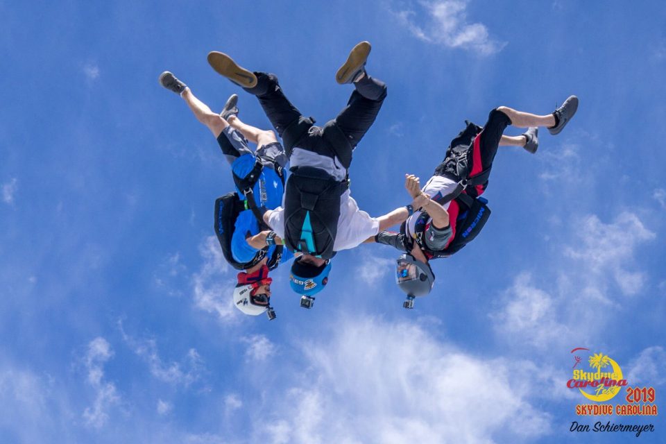 Four skydivers wearing helmets and gear form a circle mid-air against a blue sky, holding onto each other's arms and legs. Bright sunlight illuminates the scene, highlighting their colorful attire and equipment.