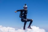 A skydiver in a black suit and helmet floats above white clouds with a blue sky as a backdrop, captured in mid-air. The diver's arms are bent at the elbows, and they are wearing sunglasses and sneakers.