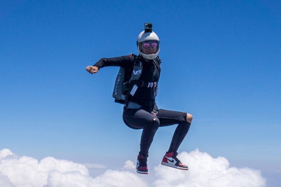 A skydiver in a black suit and helmet floats above white clouds with a blue sky as a backdrop, captured in mid-air. The diver's arms are bent at the elbows, and they are wearing sunglasses and sneakers.