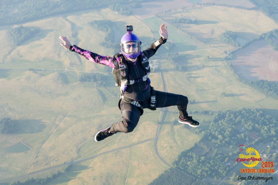 A person in a black jumpsuit and helmet skydives with arms spread wide. They wear a camera on their helmet and have a clear visor. Below, expansive green fields and winding roads are visible. The image has logos for Skydive Carolina and a date from 2019.