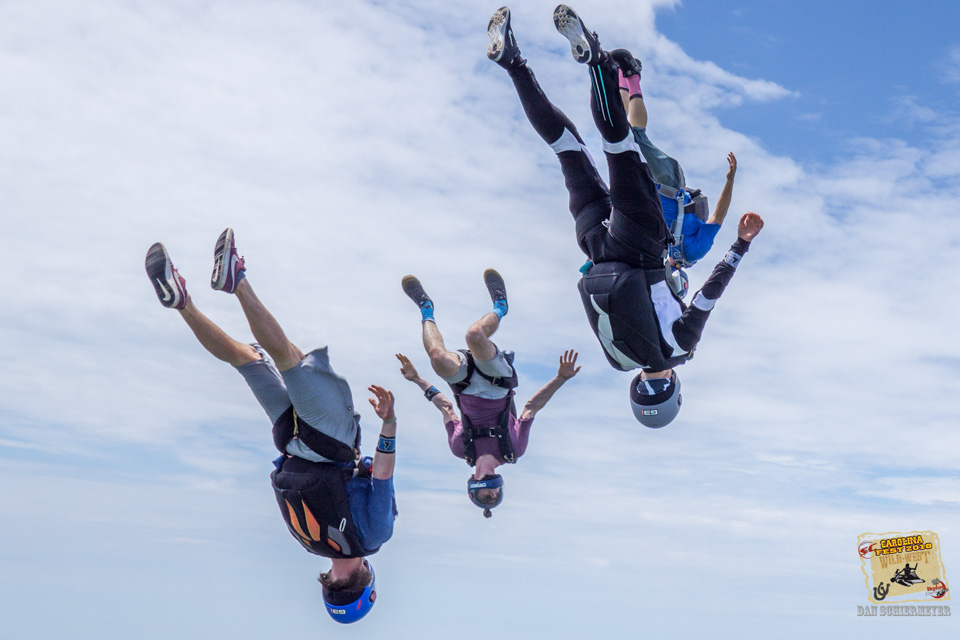 Four skydivers in mid-air perform a synchronized head-down formation against a backdrop of a clear sky. They wear helmets and jumpsuits, showcasing teamwork and skill in an energetic and coordinated dive.