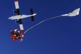 A tandem skydiving pair free-falls beneath a small plane against a clear blue sky. A parachute deployment bag trails behind them, and both individuals are wearing helmets and skydiving suits.