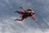 Two people tandem skydiving through partly cloudy skies, with one person in a red jumpsuit and black harness, and the other securely attached behind. They are high above a landscape of fields and forests, enjoying the free fall.