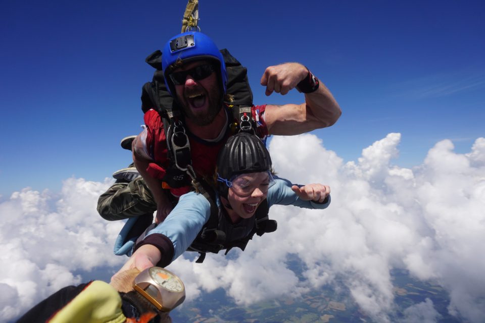 A tandem skydiving pair descends through a clear blue sky with fluffy clouds below. The instructor wears a blue helmet and flexes his arm, while the passenger wears a black helmet and shows an excited expression.