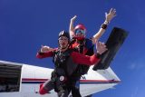 Two people tandem skydiving against a clear blue sky. They are in mid-air, just after jumping from an airplane, wearing helmets and goggles. One person is positioned behind the other, both smiling with arms outstretched.