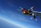 Two people tandem skydiving, jumping from an airplane. The sky is clear with a few clouds, and the horizon is visible in the background. Both wear parachute gear, and the instructor is guiding the descent.