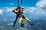 Two people skydiving tandem style, floating in the sky with green land and clouds below. The person in front wears a yellow and black jumpsuit and is smiling with arms raised, while the one behind is in a blue and red jumpsuit.