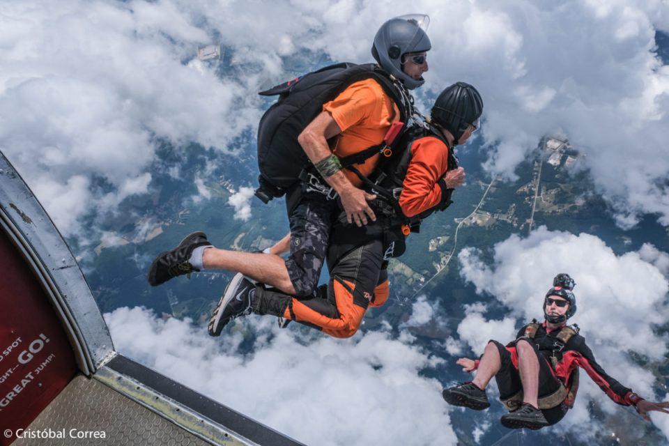 Two tandem skydivers in freefall, wearing helmets and harnesses, surrounded by clouds. Another skydiver floats nearby, capturing the scene. Below, a landscape of forests and fields is visible.