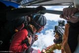 Two skydivers prepare to jump from a plane. One wears a red jumpsuit, the other an orange one with a camera mounted on the helmet. They hold onto a bar, surrounded by clouds in the sky.