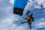 Two people tandem skydiving with a blue parachute against a cloudy sky, smiling as they descend. The person in front is wearing a yellow jumpsuit, and their shoes are visible.