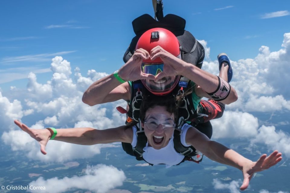 Two people tandem skydiving against a backdrop of blue sky and clouds. The instructor wears a red helmet and makes a heart shape with hands, while the woman in front smiles with arms outstretched.