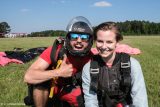 Two skydivers in gear pose on a grassy field with parachutes behind them. The person on the left gives a thumbs up, smiling widely, while the person on the right smiles gently. Clear blue sky and trees in the background.