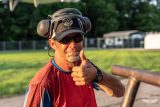 A man in sunglasses and a red shirt gives a thumbs up. He's wearing black headphones and a black cap with a logo. The background shows a grassy area and a fence with a building. The lighting suggests it's early morning or late afternoon.