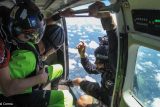 Three skydivers in a small aircraft are preparing to jump. Two are seated inside in bright gear, while one is holding onto the doorframe, looking outside. The sky is clear with scattered clouds visible below.